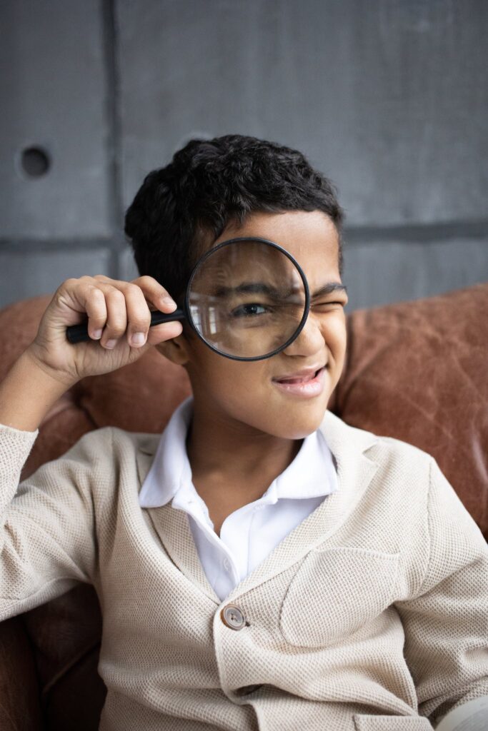 Curious  schoolboy wearing white shirt with cardigan sitting on comfortable couch and looking through magnifying glass at camera indoors