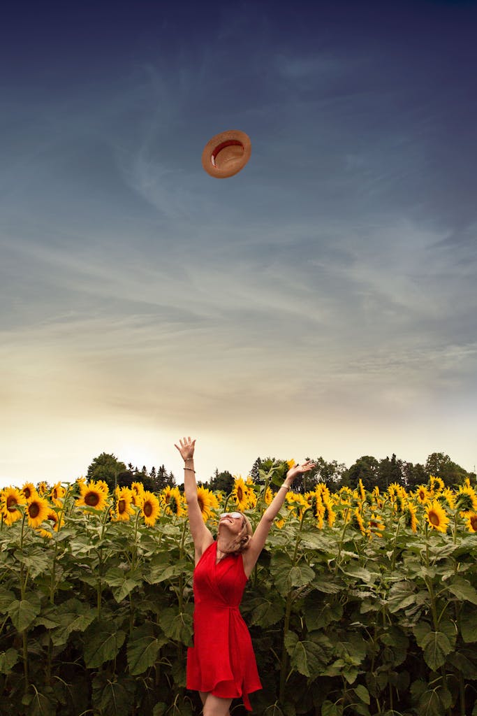 A carefree woman wearing a red dress and sunglasses joyfully throws her straw hat in a vibrant sunflower field.