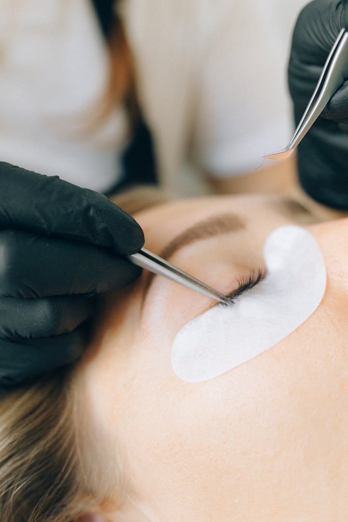 Black-gloved technician applying eyelash extensions to a woman's eye in a beauty salon setting.